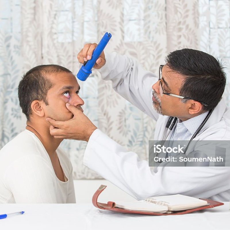 istockphoto-483852540-1024×1024 Mature Eye specialist examining a young patient, at his clinic. The doctor is looking inside the eyes of his patient with light from a torch.